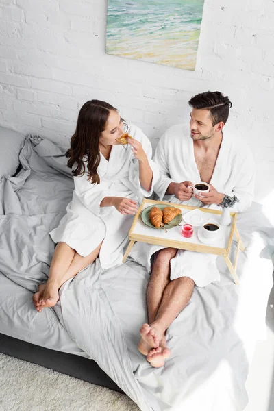 Красива дівчина і хлопець в халатах, які мають круасани і каву на сніданок у спальні — Stock Photo