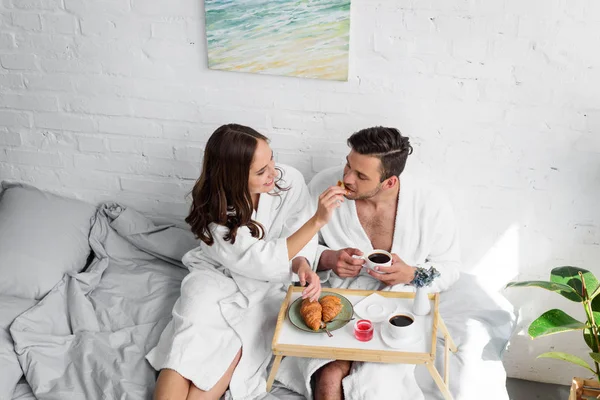Hermosa pareja joven en albornoces desayunando en la cama juntos - foto de stock