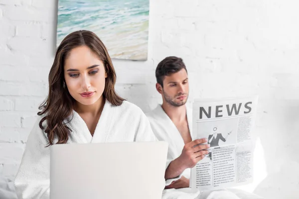 Bella fidanzata utilizzando il computer portatile mentre fidanzato leggendo giornale a letto — Foto stock