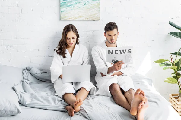 Pareja joven en albornoces sentados en la cama con ordenador portátil y periódico - foto de stock