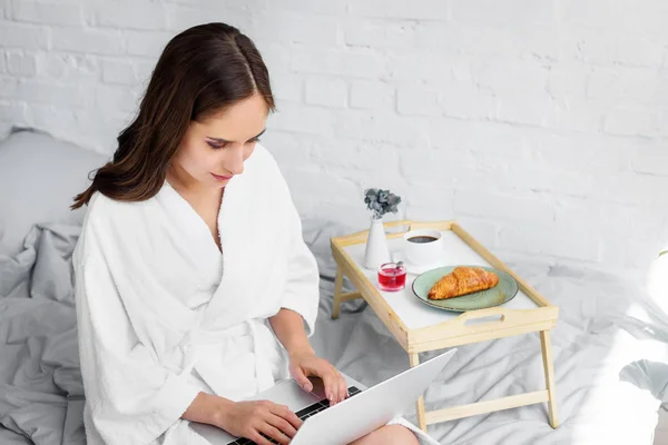 Приваблива молода жінка в білому халаті використовує ноутбук під час сніданку в спальні — стокове фото