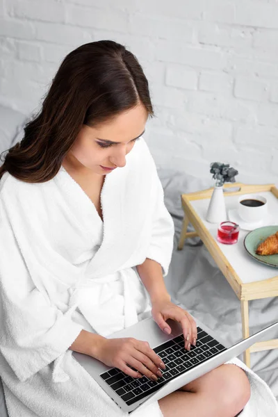 Hermosa mujer en albornoz blanco utilizando el ordenador portátil durante el desayuno en la cama - foto de stock