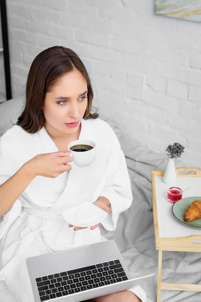 Hermosa mujer en albornoz blanco con portátil bebiendo café para el desayuno - foto de stock