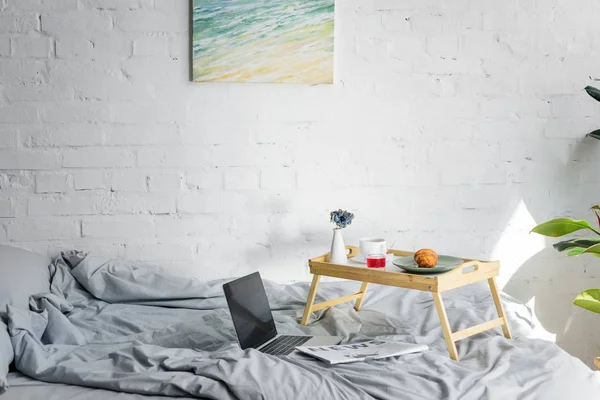 Portátil y desayuno con croissant y café en bandeja en el dormitorio con pintura en la pared - foto de stock