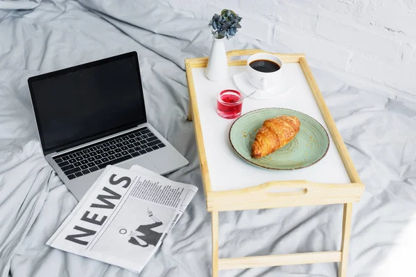 Ordinateur portable, journal et petit déjeuner avec croissant et café sur plateau au lit le matin — Photo de stock