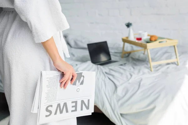 Partial view of woman with newspaper standing near bed with breakfast and laptop — Stock Photo