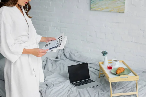 Cropped view of woman in white bathrobe reading newspaper in bedroom with laptop and breakfast on tray — Stock Photo