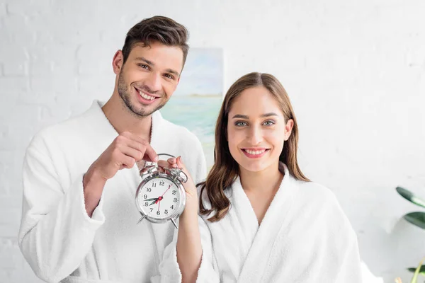 Couple souriant en peignoirs blancs tenant réveil et debout dans la chambre le matin — Photo de stock