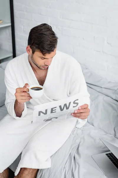 Giovane in accappatoio seduto sul letto, leggendo giornali e tenendo in mano una tazza di caffè — Foto stock