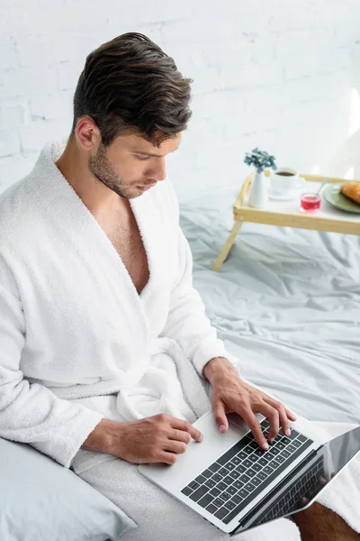 Hombre joven en albornoz sentado en la cama y el uso de ordenador portátil - foto de stock