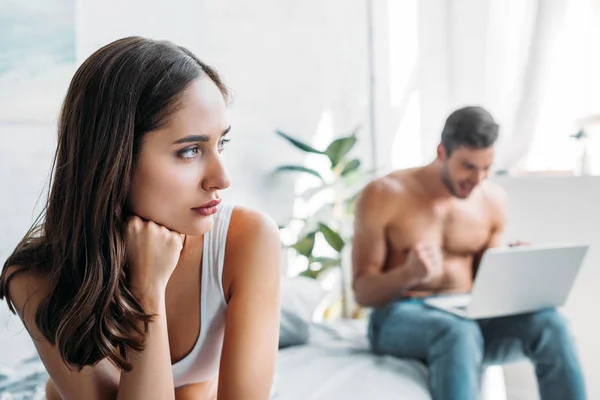 Irritated girlfriend sitting on bed and looking away while boyfriend using laptop in bedroom — Stock Photo