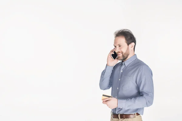 Seitenansicht eines bärtigen Mannes mit Kreditkarte in der Hand, der auf einem Smartphone isoliert auf weiß spricht — Stockfoto