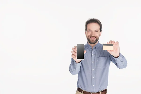 Retrato de homem barbudo sorridente mostrando smartphone com tela em branco e cartão de crédito em mãos isoladas em branco — Fotografia de Stock