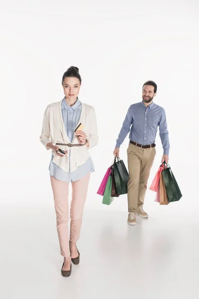 Женщина с кредитной картой и смартфоном смотрит в камеру, в то время как муж несет сумки изолированы на белом — стоковое фото