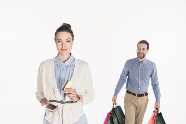 Mujer con tarjeta de crédito y teléfono inteligente mirando a la cámara mientras el marido lleva bolsas de compras aisladas en blanco - foto de stock
