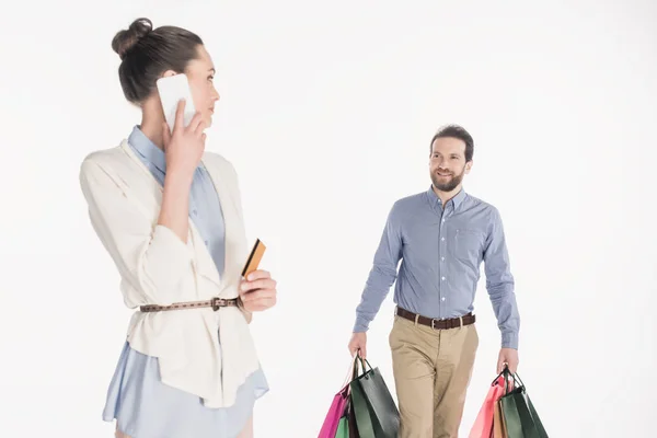 Frau mit Kreditkarte spricht auf Smartphone, während Mann Einkaufstüten auf weißem Grund trägt — Stockfoto