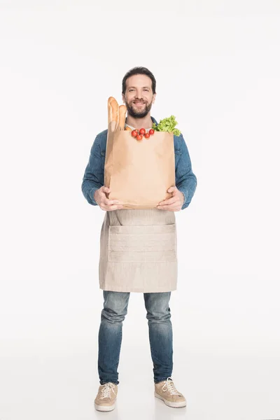 Hombre barbudo en delantal con paquete de papel lleno de alimentos aislados en blanco - foto de stock