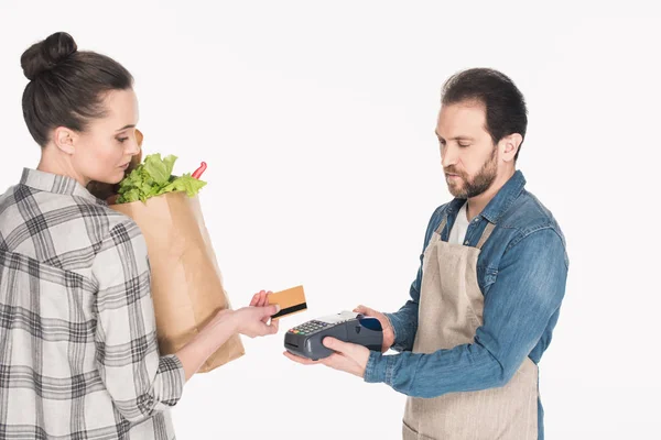 Vista lateral de la mujer con el paquete de papel con la comida que da tarjeta de crédito al ayudante de la tienda con el lector de la tarjeta aislada en blanco - foto de stock