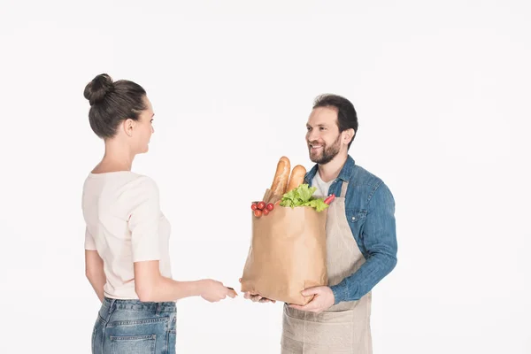 Side view of woman giving credit card to shop assistant in apron with paper package full of food isolated on white — Stock Photo