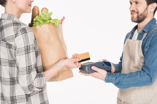 Vista parcial de la mujer con el paquete de papel con la comida que da tarjeta de crédito al asistente de la tienda con el lector de tarjetas aislado en blanco - foto de stock