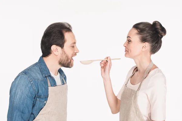 Seitenansicht einer lächelnden Frau mit Kochlöffel, die vorgibt, Ehemann isoliert auf Weiß zu ernähren — Stockfoto