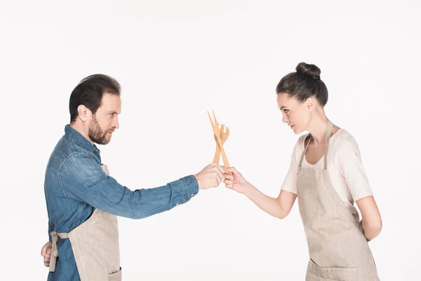 Side view of couple in aprons pretending to fight with wooden kitchen utensils isolated on white — Stock Photo