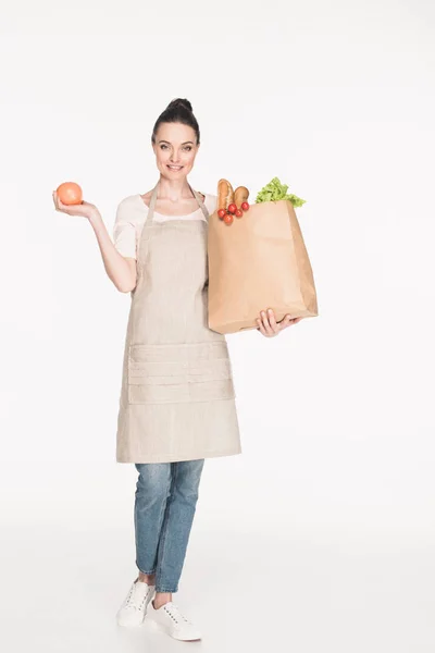 Donna sorridente in grembiule contenente pacchetto di carta con generi alimentari isolati su bianco — Foto stock