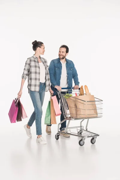 Casal com sacos de compras e embalagens de papel cheias de alimentos no carrinho de compras isolado em branco — Fotografia de Stock