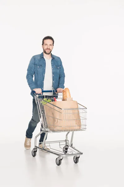 Hombre sonriente con carrito de compras lleno de paquetes de papel con supermercado aislado en blanco - foto de stock