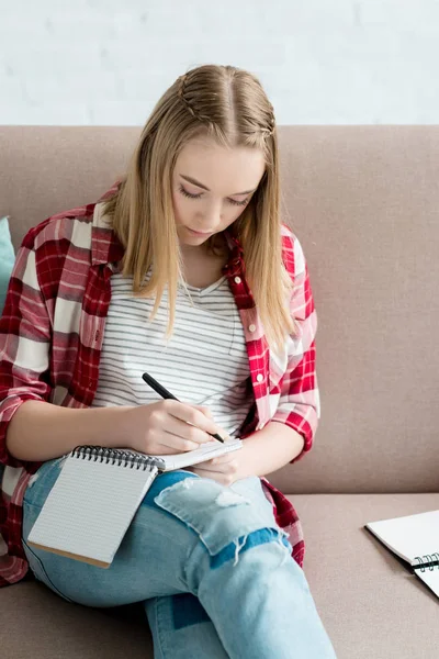 Teenager-Studentin schreibt in Notizbuch, während sie auf dem Sofa sitzt und Hausaufgaben macht — Stockfoto