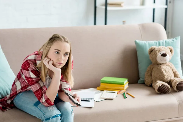 Annoiato adolescente studente ragazza con telecomando guardando la tv mentre seduto sul divano con libro e note — Foto stock