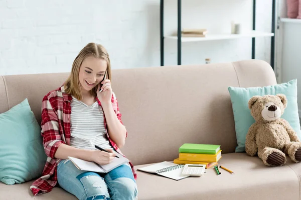 Teen student girl doing homework on couch and talking by phone — Stock Photo