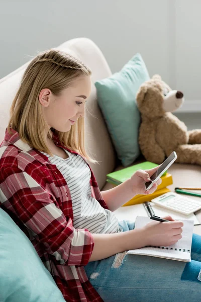 Adolescent étudiant fille faire des devoirs sur canapé et en utilisant smartphone — Photo de stock