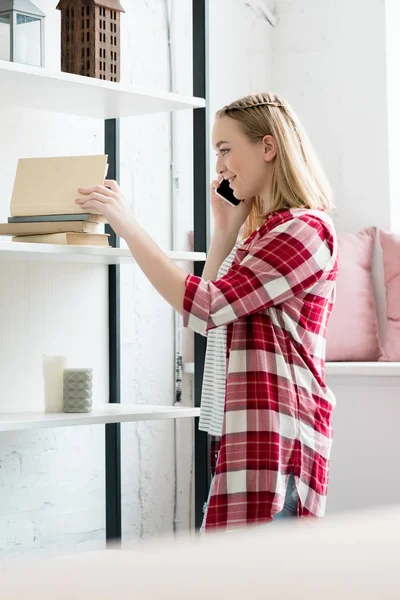 Teen student girl talking by phone and taking book from shelf — Stock Photo