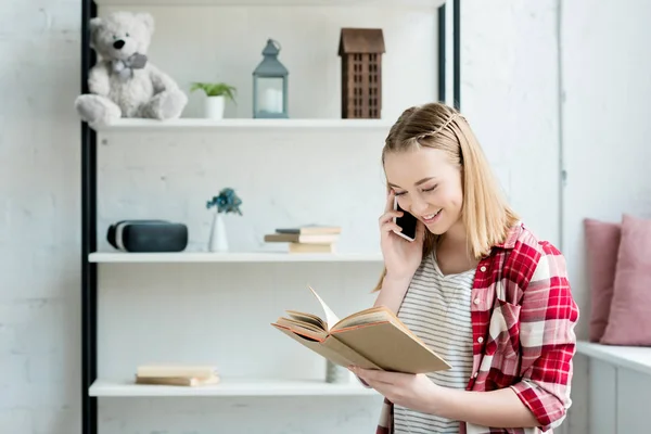 Smiling teen student girl reading book and talking by phone at home — Stock Photo