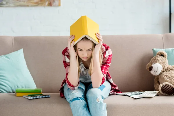 Depressed teen student girl with book on head sitting on couch at home — Stock Photo