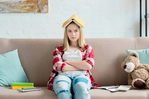 Sad teen student girl with book on head sitting on couch at home and looking at camera — Stock Photo