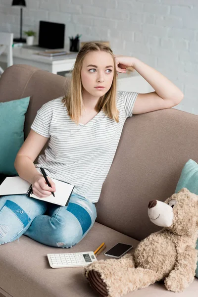 Thoughtful teen student girl with notebook sitting on couch and looking away — Stock Photo