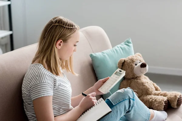 Adolescente estudiante chica escritura en cuaderno y el uso de calculadora mientras está sentado en sofá — Stock Photo