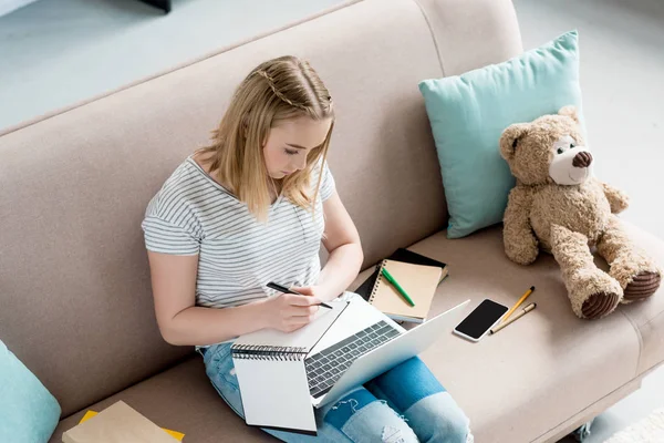 Вид под высоким углом на школьницу-подростка, выполняющую домашнюю работу сидя на диване — стоковое фото