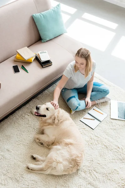 Blick aus der Vogelperspektive auf ein hübsches Teenie-Mädchen, das ihren Hund streichelt, während es Hausaufgaben macht — Stockfoto