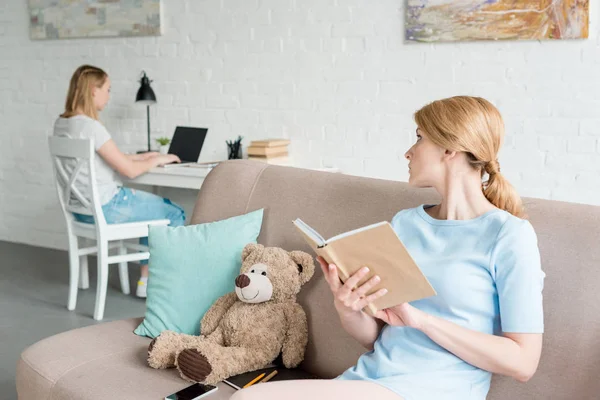 Madre leyendo libro y sentada en el sofá mientras su hija trabaja con el ordenador portátil — Stock Photo