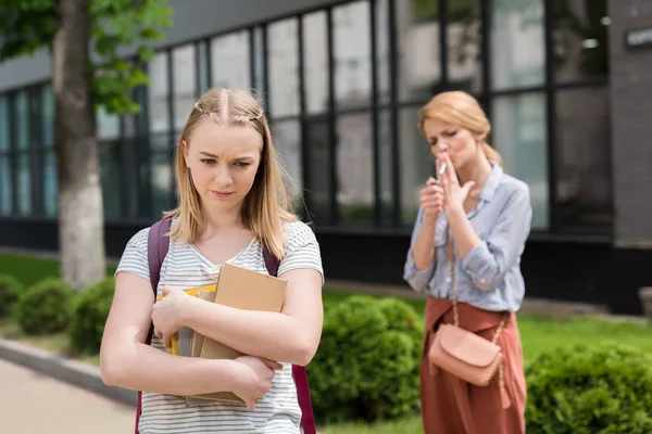 Deluso figlia adolescente con i libri guardando verso il basso mentre sua madre fumare sigaretta offuscata sullo sfondo — Foto stock