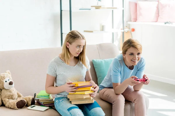 Glückliche Mutter beim Konsolenspiel, während ihre verwirrte Tochter mit einem Stapel Bücher auf der Couch sitzt — Stockfoto