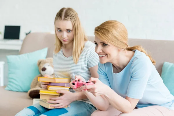 Mutter spielt Konsolenspiel, während ihre verwirrte Tochter mit einem Stapel Bücher auf der Couch sitzt — Stockfoto