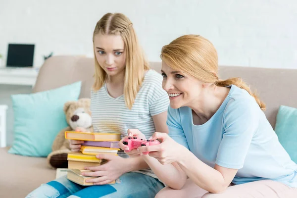 Madre giocare console gioco mentre la sua confusa figlia seduta con pila di libri sul divano — Foto stock