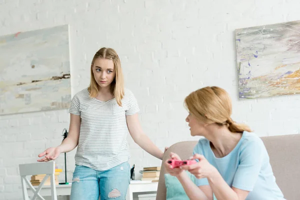 Desconcertada hija adolescente mirando a la madre mientras ella juega juego de consola - foto de stock