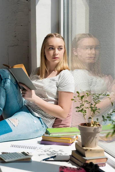 Вдумлива студентка-підліток з книгою, що сидить на підвіконні — стокове фото