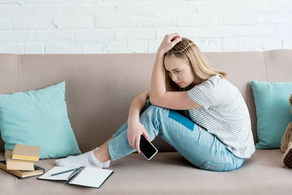Депрессивная девочка-подросток сидит на диване со смартфоном и делает домашнее задание — стоковое фото