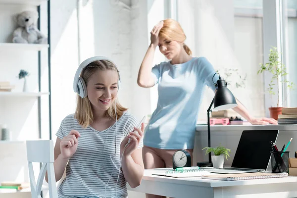 Feliz adolescente hija escuchando música mientras su decepcionada madre de pie en el fondo - foto de stock